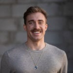 Professional portrait of a man with short brown hair and a mustache, wearing a gray sweater and a circular pendant necklace, smiling confidently in front of a blurred gray wall.