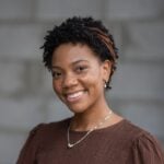 Confident woman with short curly hair, brown headband, brown top, gold earrings, and necklace smiles in front of a blurred gray wall. Featured on Hook Agency’s website.