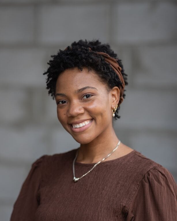 Confident woman with short curly hair, brown headband, brown top, gold earrings, and necklace smiles in front of a blurred gray wall. Featured on Hook Agency’s website.