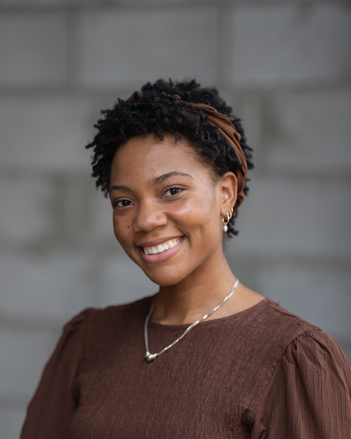 Confident woman with short curly hair, brown headband, brown top, gold earrings, and necklace smiles in front of a blurred gray wall. Featured on Hook Agency’s website.