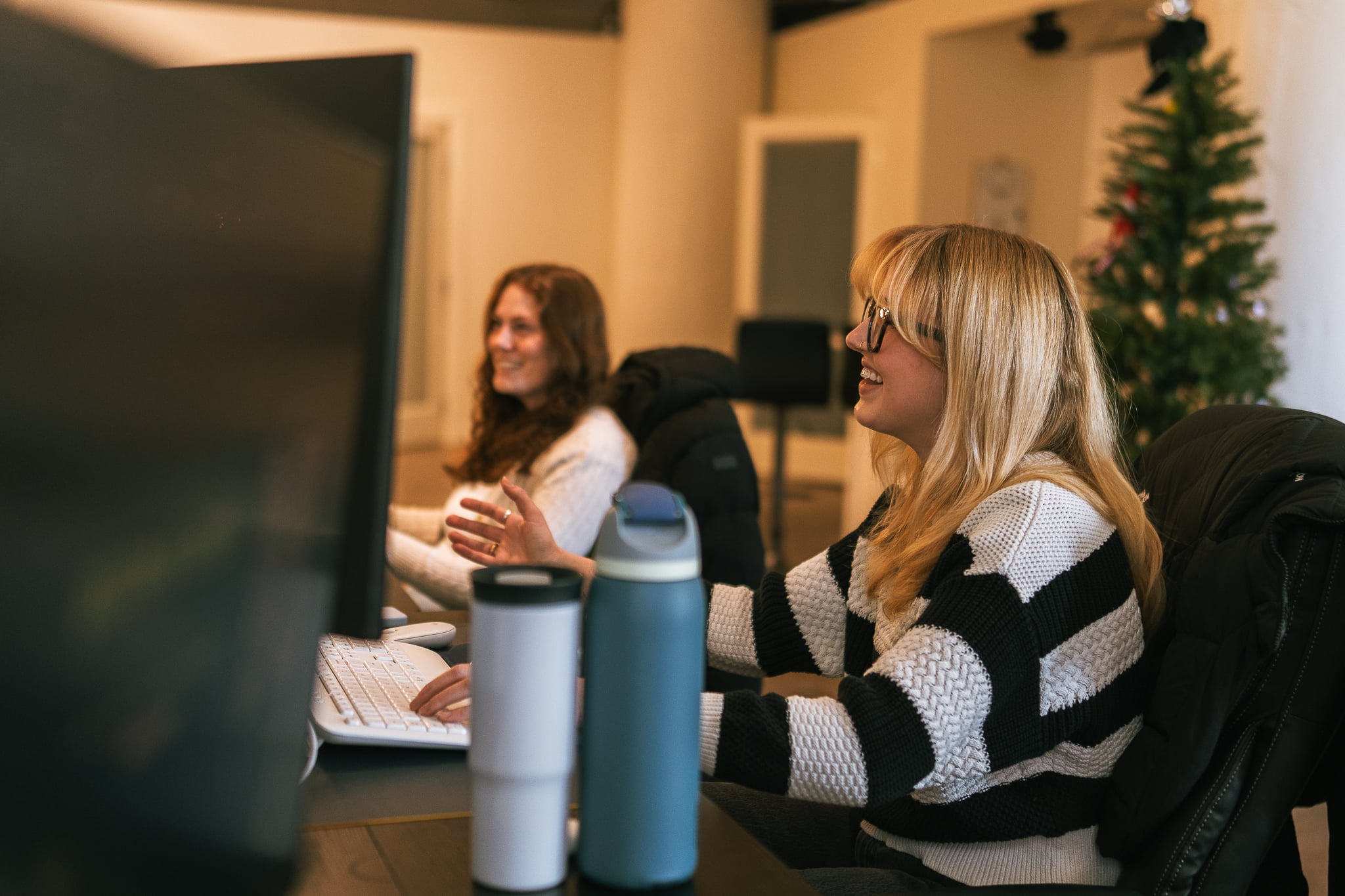 Two team members at Hook Agency work on computers in a modern office, with one smiling enthusiastically. Desks have water bottles, and a decorated Christmas tree is in the background.