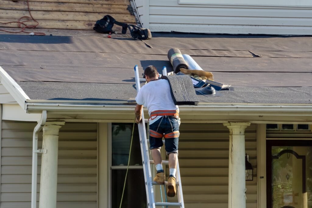 Professional roofer ascends a ladder carrying shingles while homeowners supervise; roofing materials and tools are organized on the partially underlayment-covered roof, demonstrating a safe and systematic approach to roof replacement.