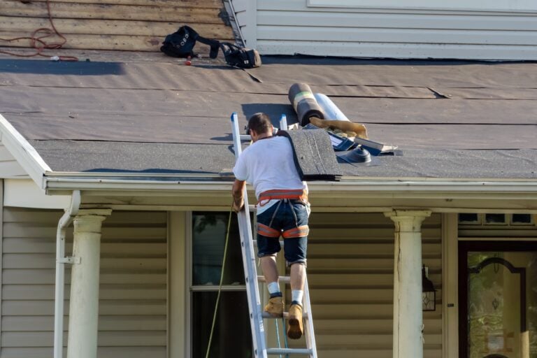 Professional roofer ascends a ladder carrying shingles while homeowners supervise; roofing materials and tools are organized on the partially underlayment-covered roof, demonstrating a safe and systematic approach to roof replacement.