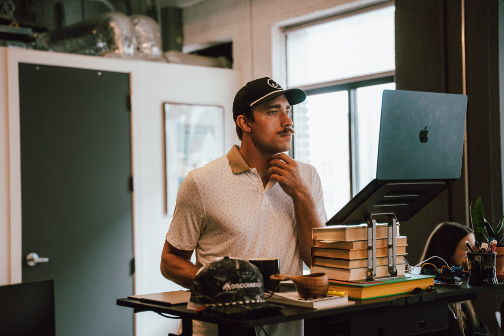 A man with a mustache and baseball cap reviews an SEO report on a laptop elevated by books.